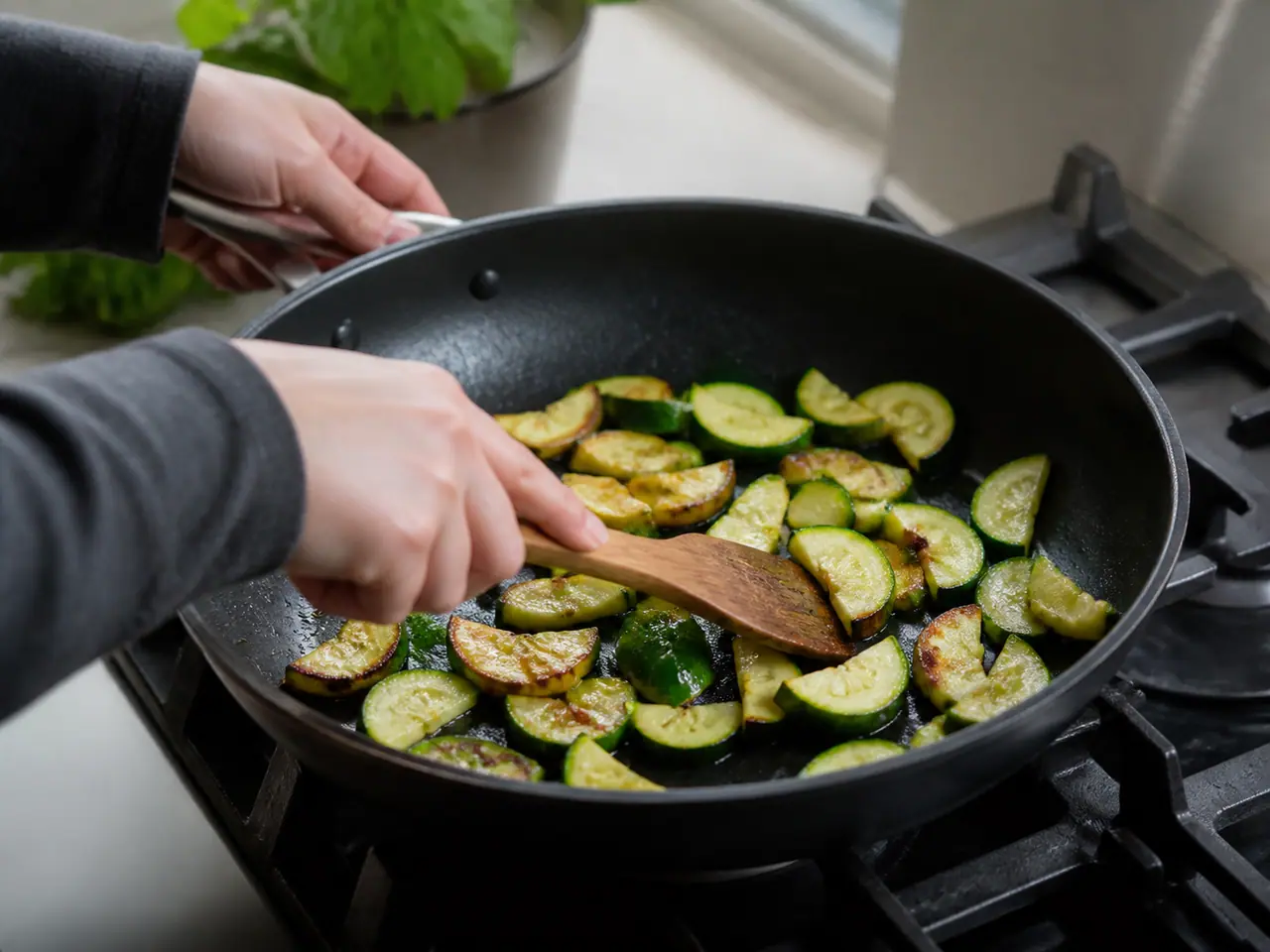 Hands turning browned courgette pieces in a wide frying pan