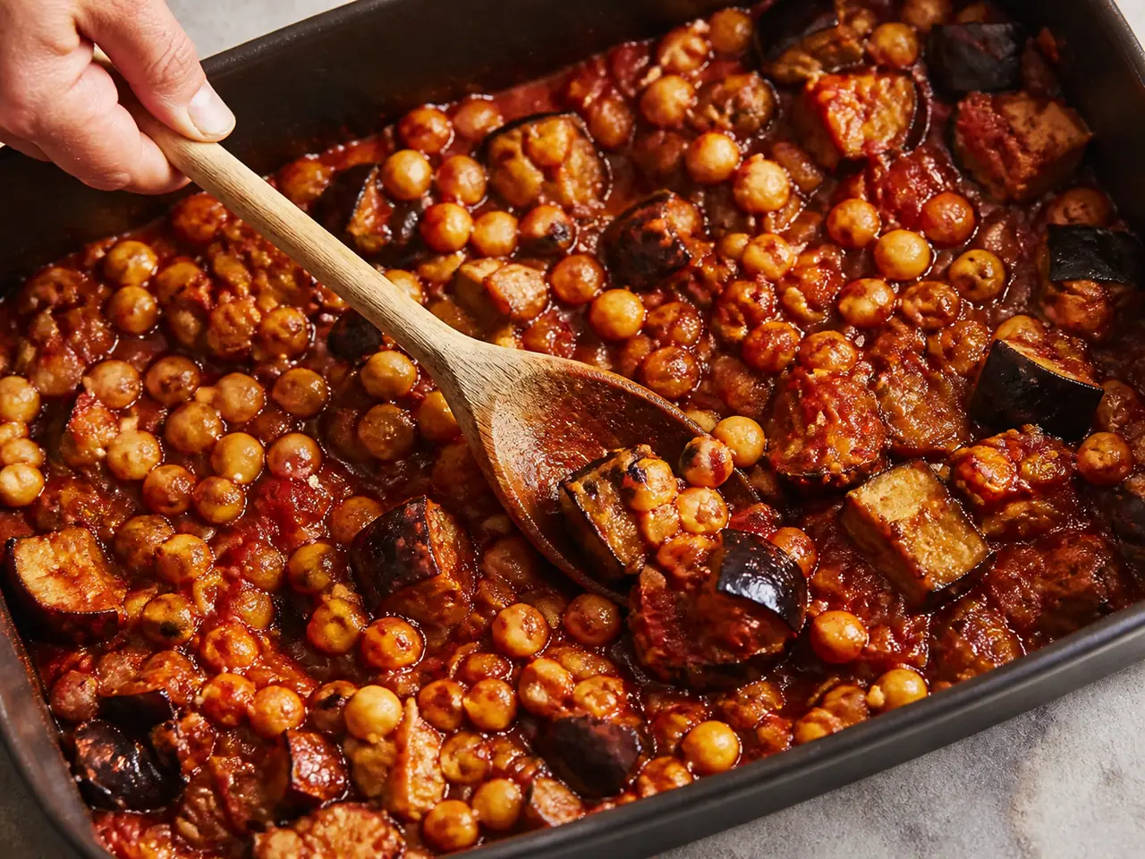 Hands stirring harissa aubergine with chickpeas for the freezer in a roasting tin