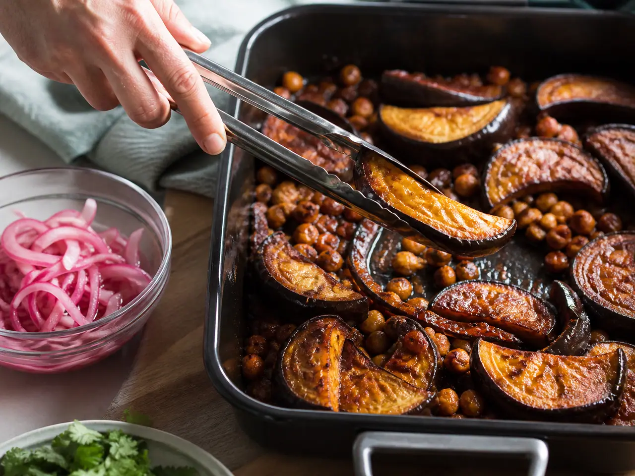 Hands stirring harissa aubergine with chickpeas with pickled onions in a roasting tin
