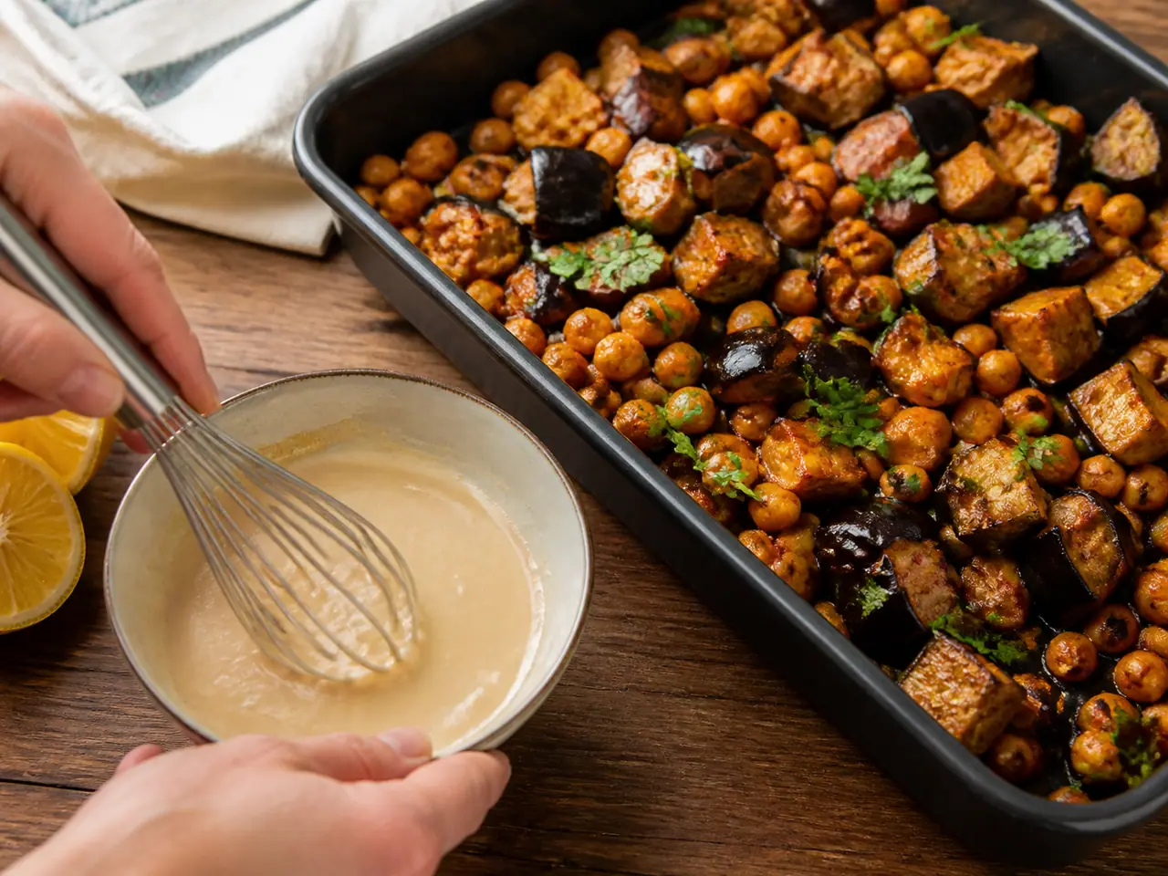 Hands stirring harissa aubergine with chickpeas with tahini dressing in a roasting tin