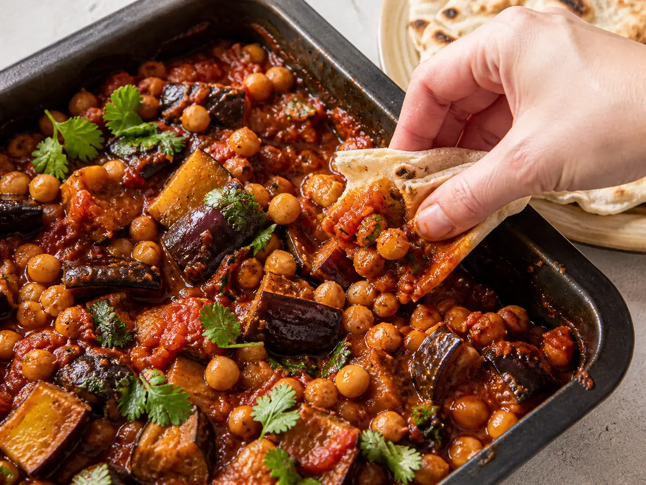 Hands stirring harissa aubergine with chickpeas with warm flatbread in a roasting tin
