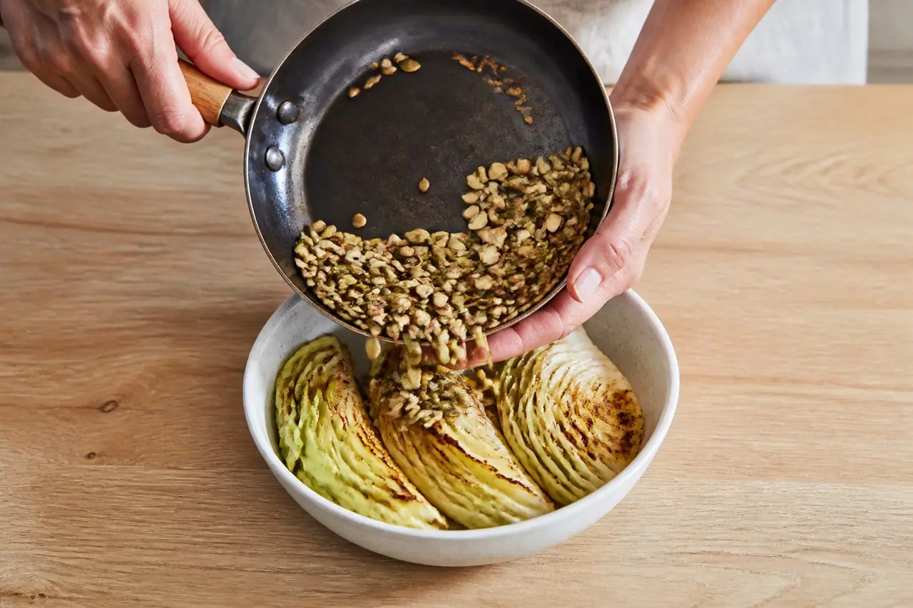 Hands tipping toasted pumpkin and sunflower seeds into a bowl beside roasted cabbage