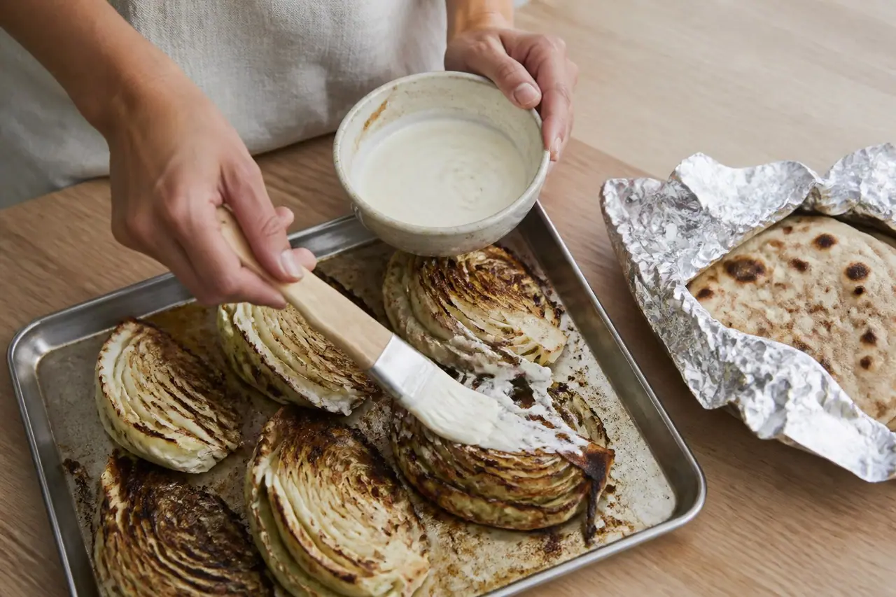 Hands brushing miso glaze over cabbage wedges before returning the tray to the oven