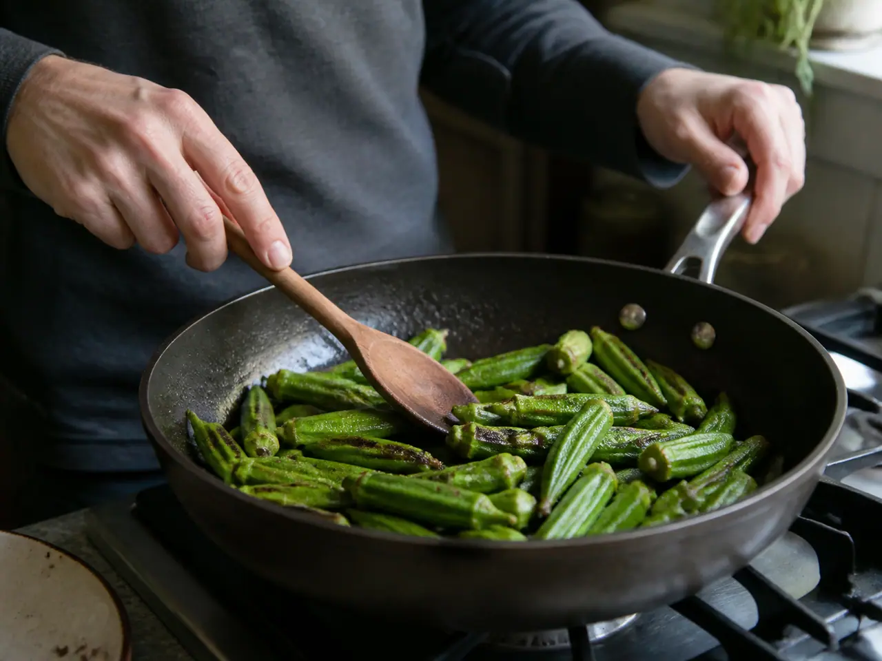 Hands turning dry-seared okra pieces in a wide frying pan