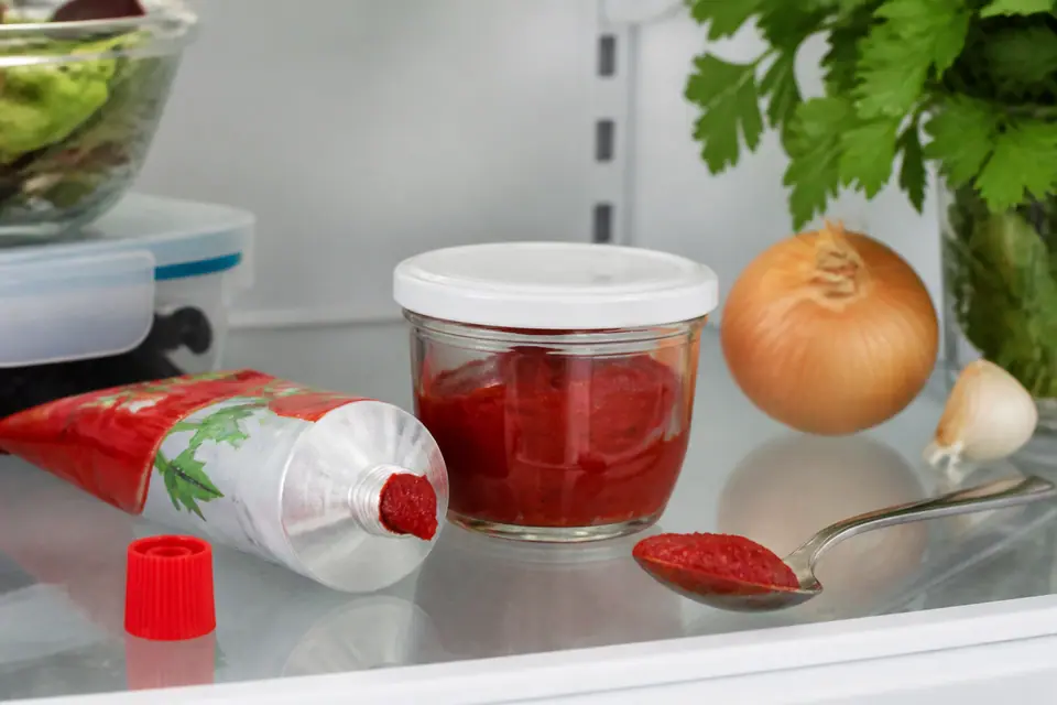 Covered container of tomato paste beside a spoon and an open tube