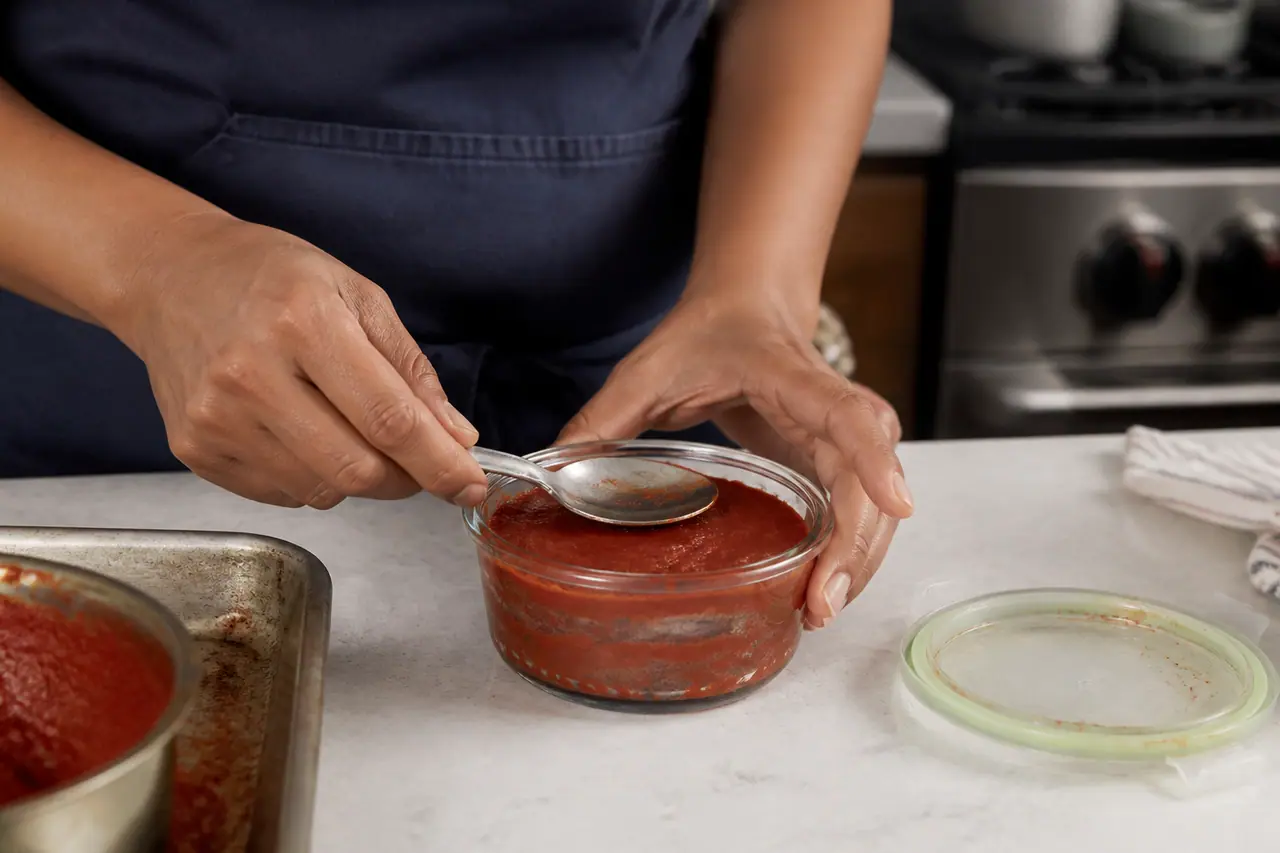 Hands smoothing tomato paste into a small covered glass container
