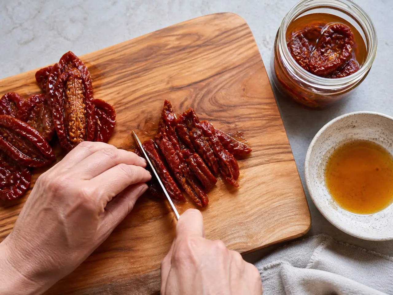 Hands slicing oil-packed sun-dried tomatoes on a wooden board