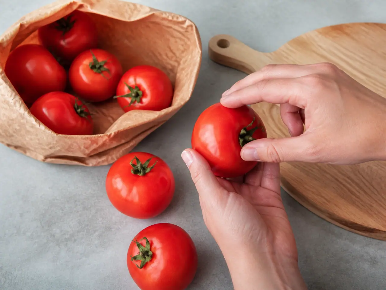 Hands checking ripe tomatoes beside a paper bag and cutting board