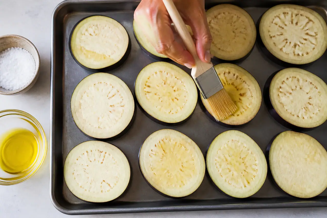 Hand brushing measured oil over aubergine slices on a tray